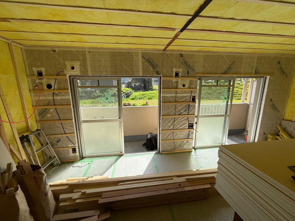 住宅リノベーションの施工風景。壁に遮熱シート「SUKATTO SHEET PREMIUM」が施工されている様子。天井の断熱材や積まれた木材、掃き出し窓が見える。